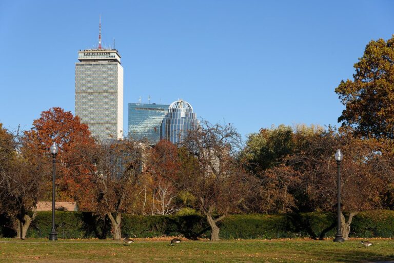 The Back Bay Fens in fall, with a lawn, lampposts and trees in orange and red in front of the Prudential Tower and the Back Bay skyline under a clear blue sky.