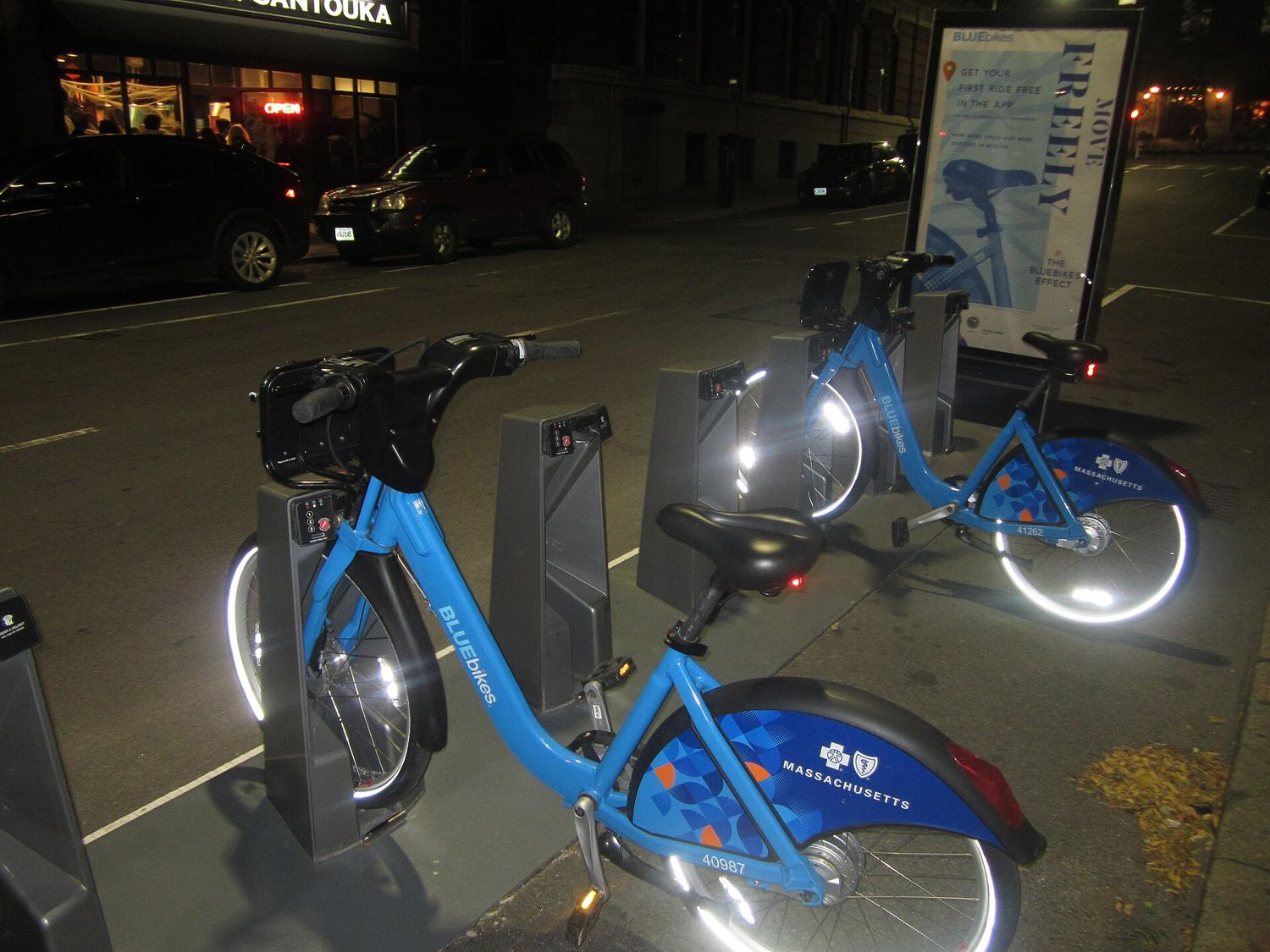 A Bluebikes docking station on a Boston street at night, with two blue bikes locked into the dock and an information kiosk beside it.