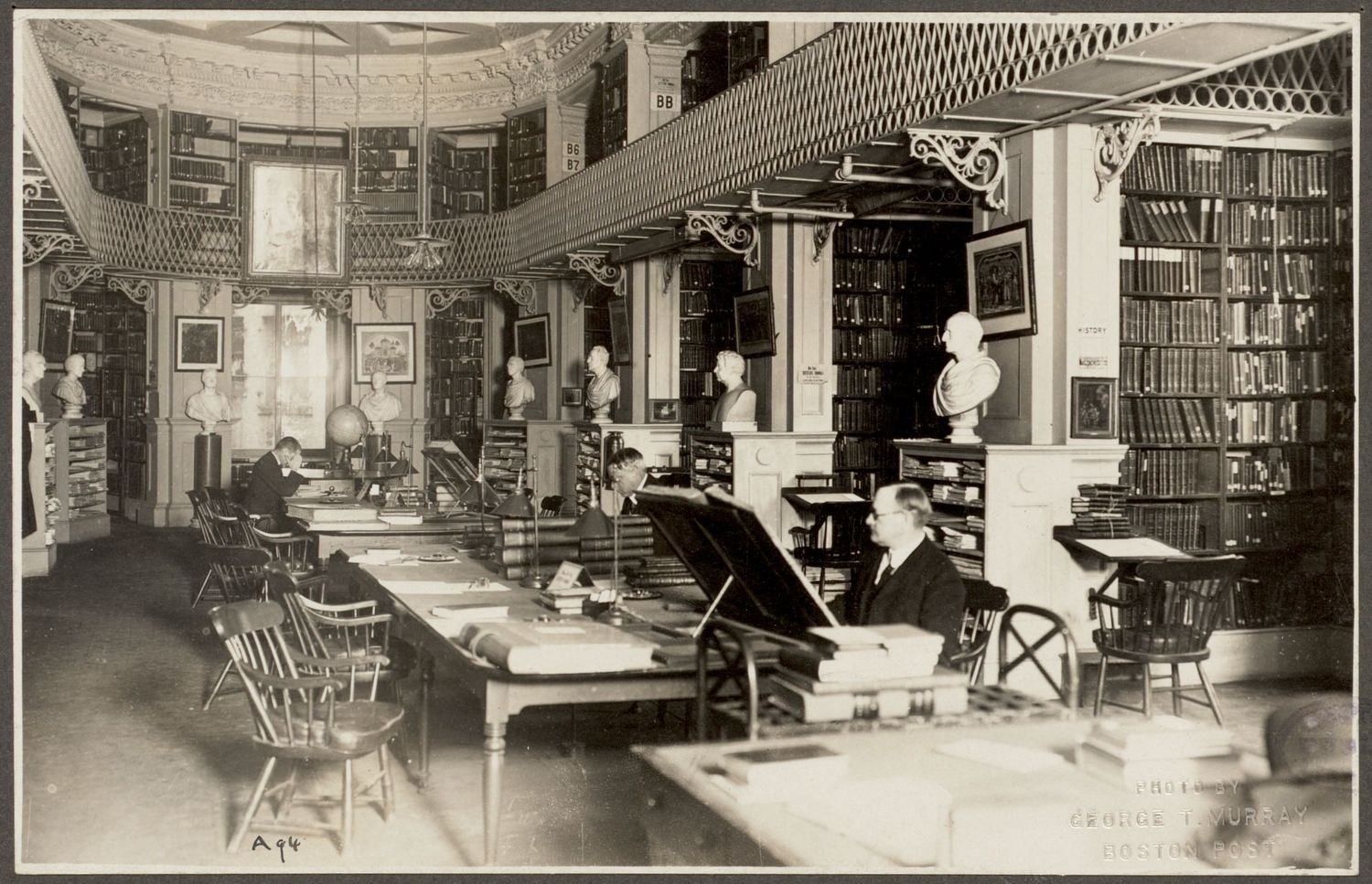An early twentieth-century photograph of a reading room at the Boston Athenaeum, with readers at long tables, tall bookcases, marble busts on pedestals and an ornate iron gallery running around the upper level.