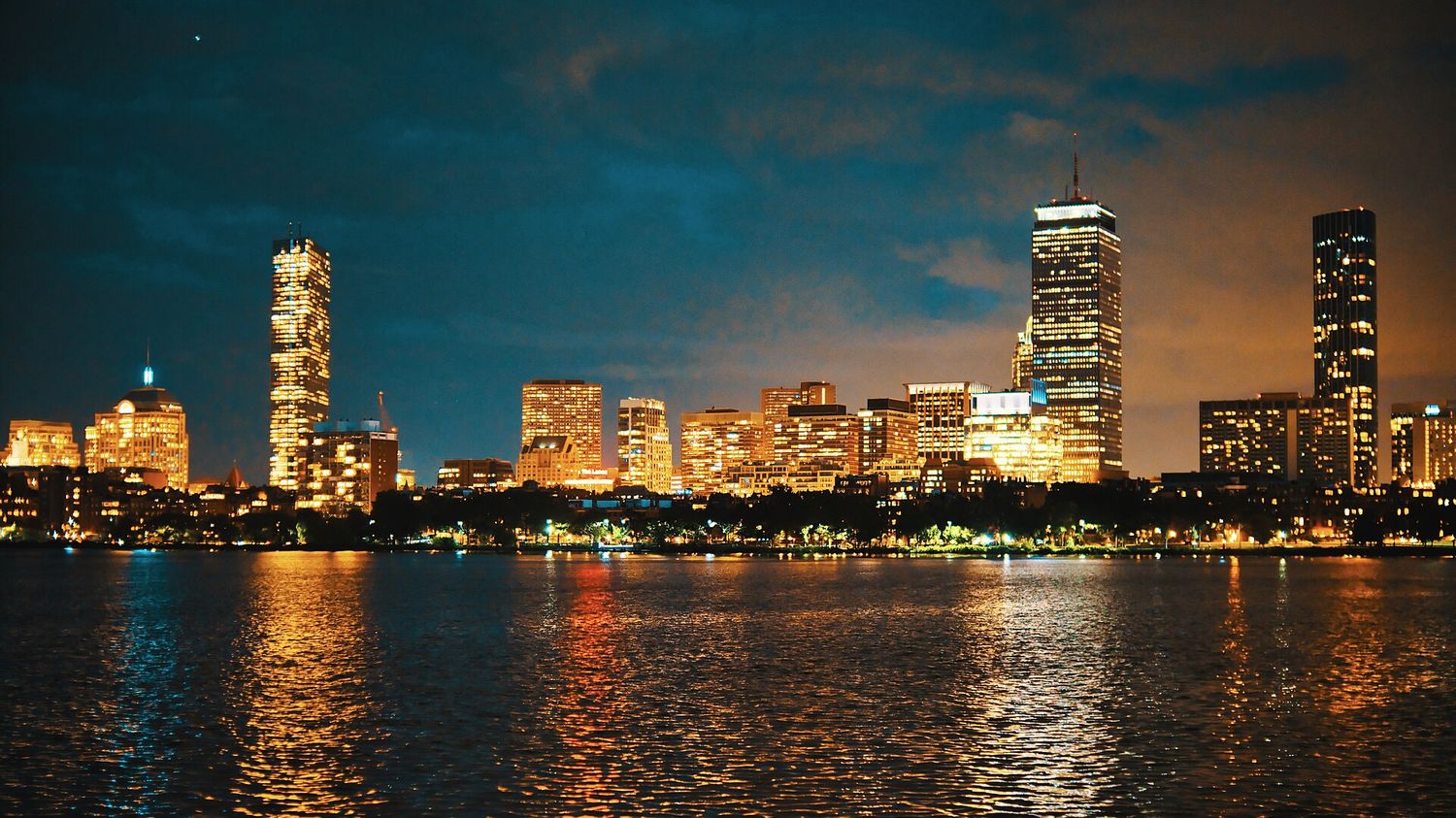 The Back Bay skyline of Boston lit up at night, reflected in the Charles River, with the Prudential Tower and 200 Clarendon standing above the lower blocks.