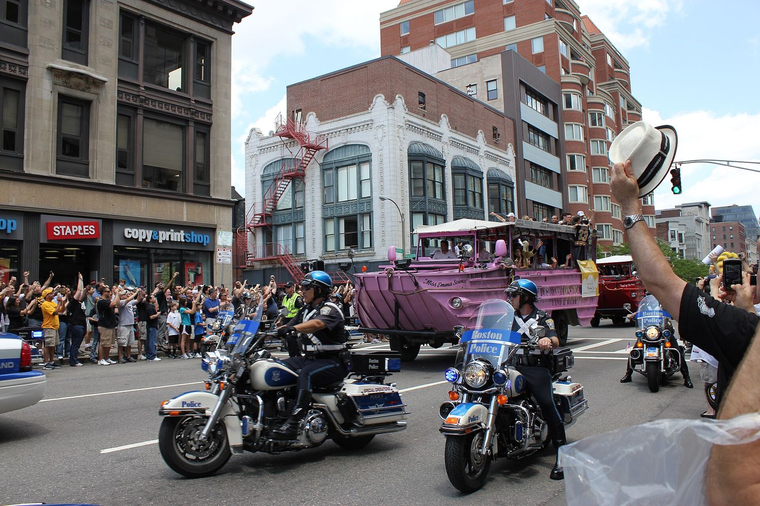 A pink Boston Duck Tours vehicle carrying Boston Bruins players through a downtown street during the 2011 Stanley Cup parade, escorted by police motorcycles, with cheering fans lining the sidewalk.