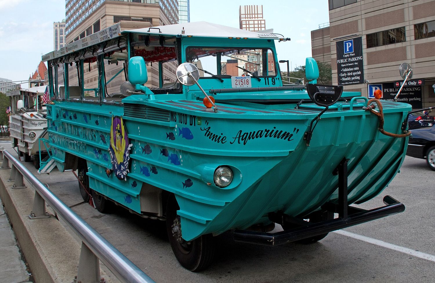 A teal Boston Duck Tours vehicle named Annie Aquarium, painted with blue fish, parked at the curb beside office towers and a Copley Place public parking sign, with a white Duck parked behind it.