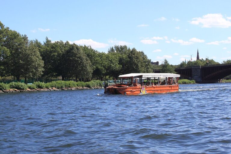 An orange Boston Duck Tours vehicle afloat on the Charles River beside a tree-lined riverbank, with a stone-arched bridge and a church spire in the distance.