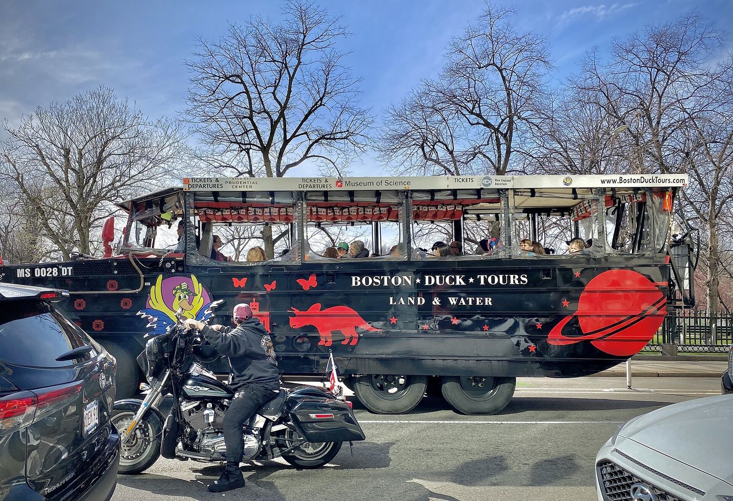 A black Boston Duck Tours amphibious vehicle painted with a red planet and a dinosaur, carrying passengers under a canopy along a city street lined with bare trees, with a motorcyclist waiting alongside.