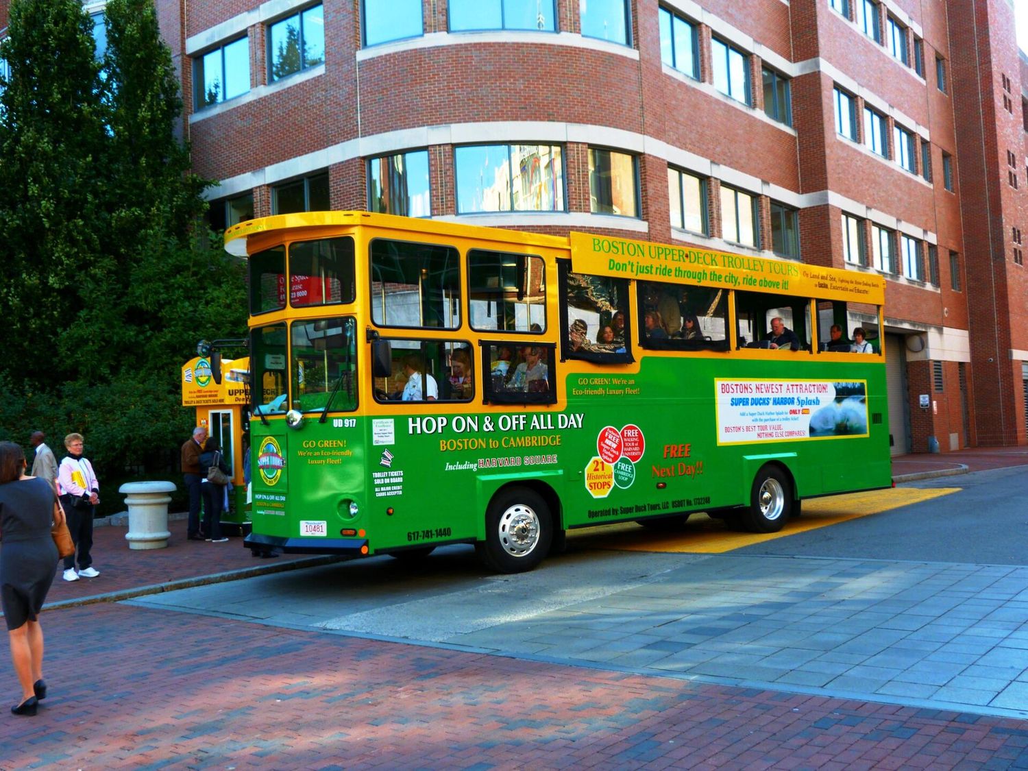 A green and yellow Boston Upper Deck hop-on hop-off trolley loading passengers beside a brick building, with a ticket kiosk on the sidewalk.
