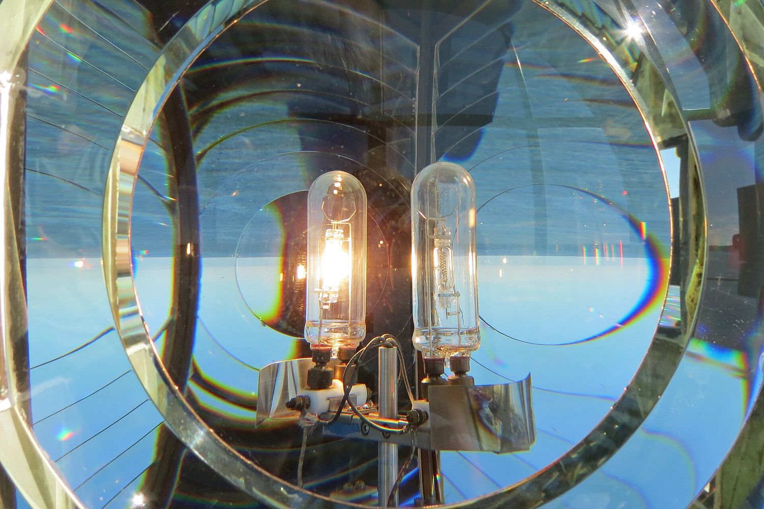 Close-up of the glass Fresnel lens inside Boston Light on Little Brewster Island, with two lit bulbs at its center and concentric prism rings around them.