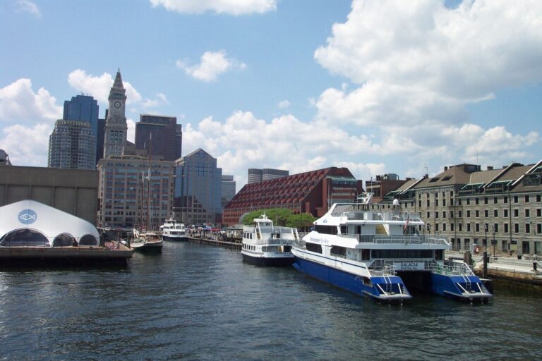 Passenger ferries moored at Long Wharf on Boston's waterfront, with the Custom House clock tower and downtown towers behind and a white tent on the Aquarium's wharf to the left.