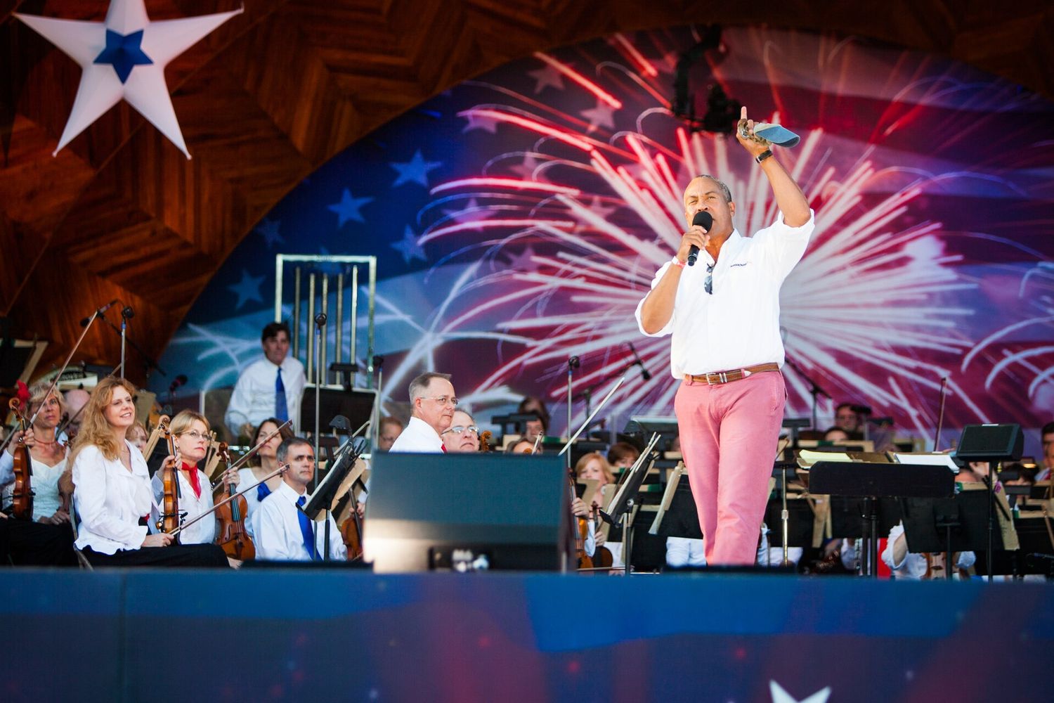 The Boston Pops orchestra on the Hatch Shell stage on the Charles River Esplanade during a Fourth of July concert, with a fireworks-themed backdrop behind the players.