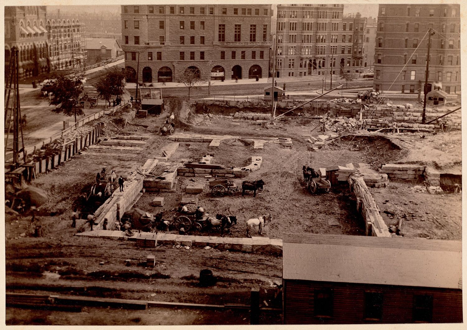 A sepia photograph from 1888 showing the foundations of the Boston Public Library under construction, with horse-drawn carts, stone blocks and workmen on the site and Back Bay buildings behind.