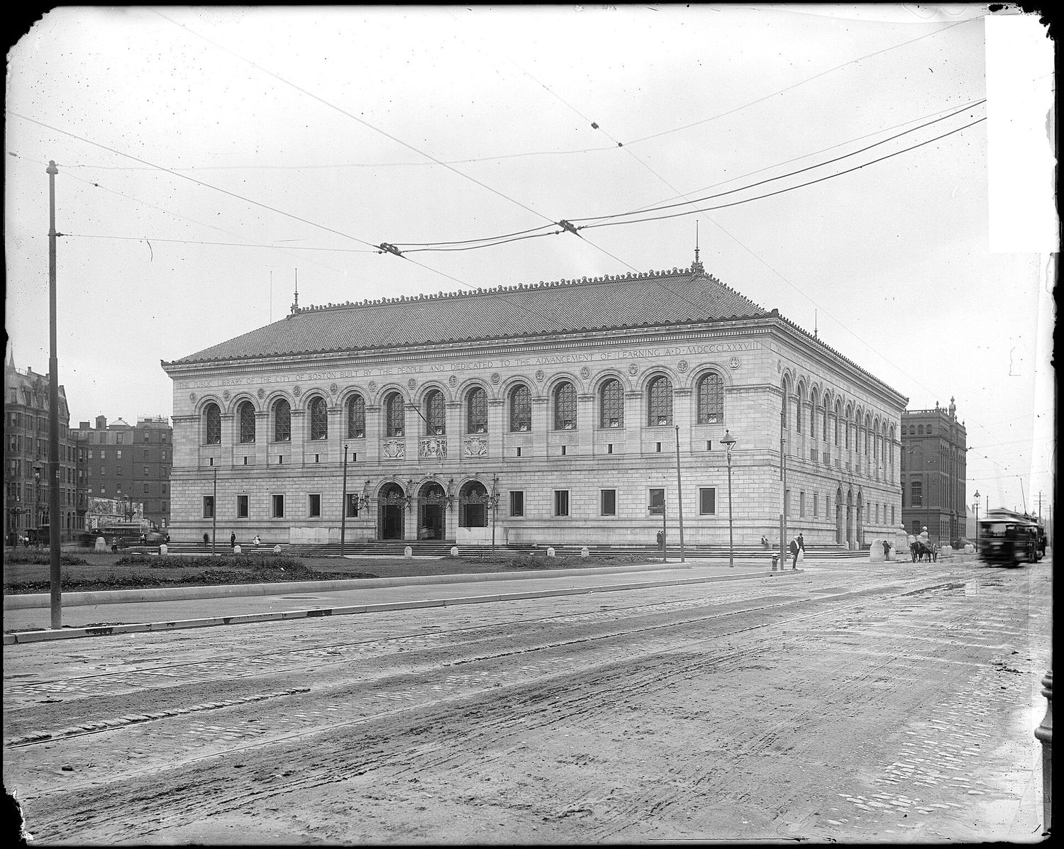 An early black-and-white photograph of the Boston Public Library's McKim Building soon after completion, with a streetcar and horse-drawn carriage on the unpaved street in front and overhead trolley wires.
