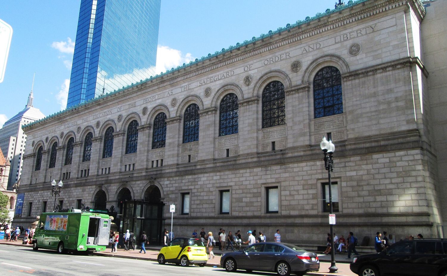 The long granite side of the McKim Building of the Boston Public Library, with a row of tall arched windows and a carved inscription reading The Commonwealth requires the education of the people as the safeguard of order and liberty, and the glass tower of 200 Clarendon rising behind.