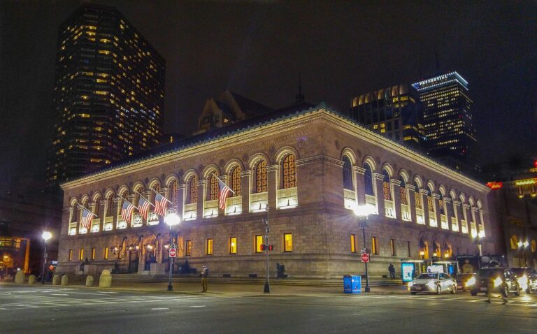 The Boston Public Library's McKim Building lit up at night on Copley Square, its arched windows glowing and American flags hanging from the facade, with office towers and the Prudential Tower behind.