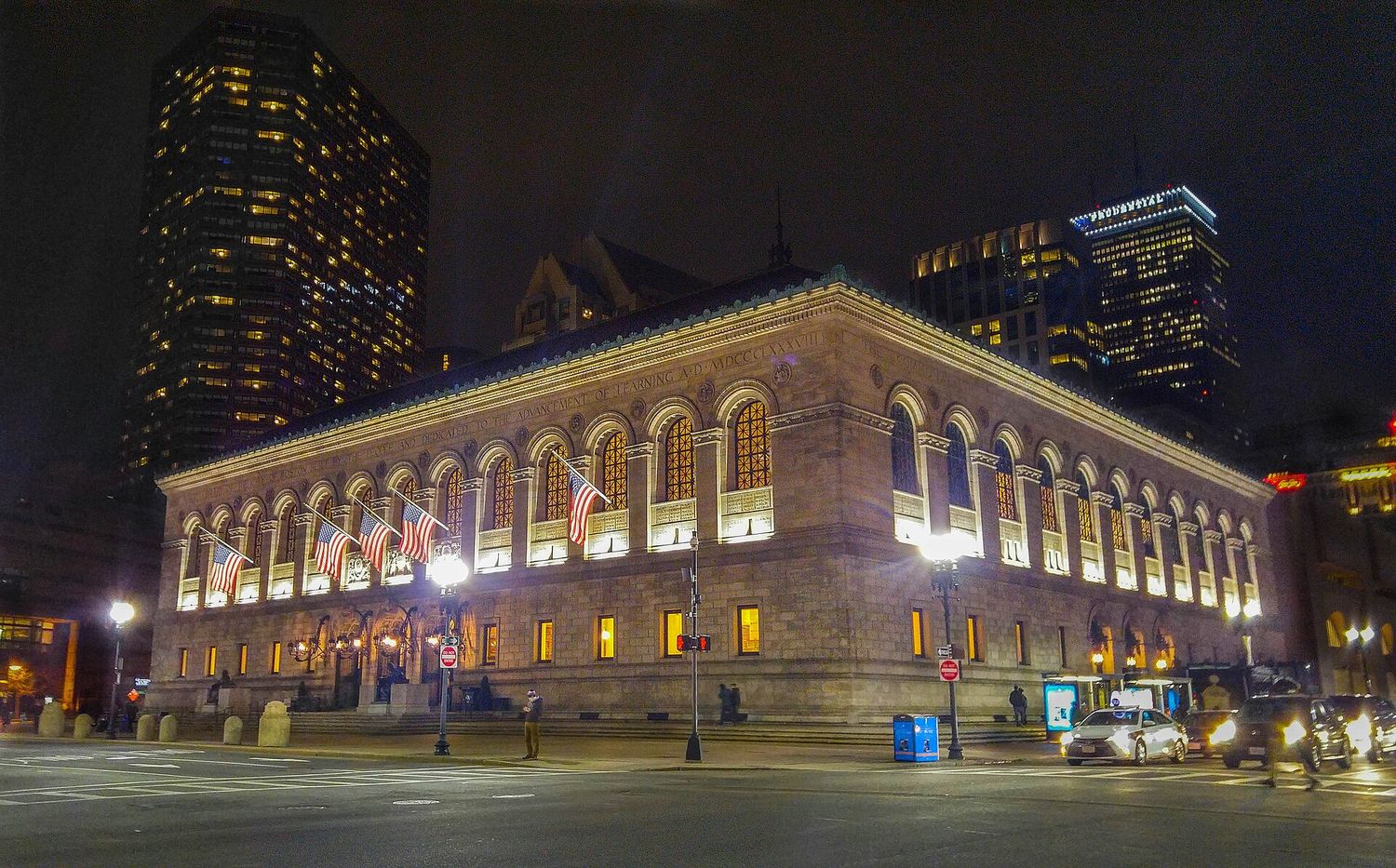 The Boston Public Library's McKim Building lit up at night on Copley Square, its arched windows glowing and American flags hanging from the facade, with office towers and the Prudential Tower behind.