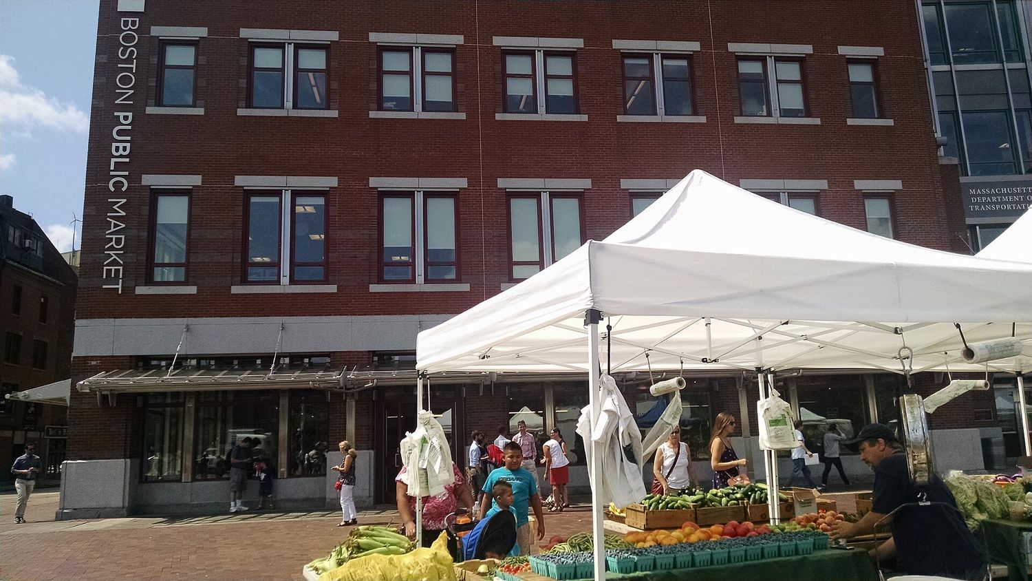 Farmers' market stalls under white canopies on the plaza outside the brick Boston Public Market building, with crates of corn, tomatoes and berries in the foreground.