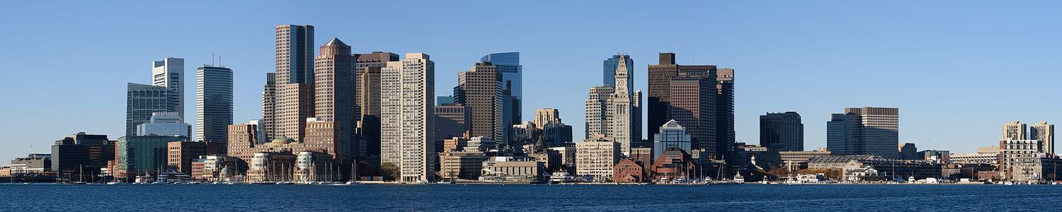 A wide panorama of the downtown Boston skyline across the harbor from East Boston, with the Custom House clock tower in the middle and wharves along the waterfront under a clear blue sky.