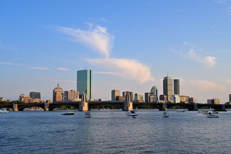 The Back Bay skyline, with 200 Clarendon and the Prudential Tower, rising behind the arches of the Longfellow Bridge across the Charles River on a summer evening, with small boats moored in the foreground.