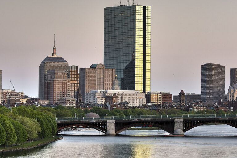 The Back Bay skyline at sunset from the roof of the Museum of Science, with the glass tower of 200 Clarendon glowing gold above the Longfellow Bridge and the tree-lined bank of the Charles River.
