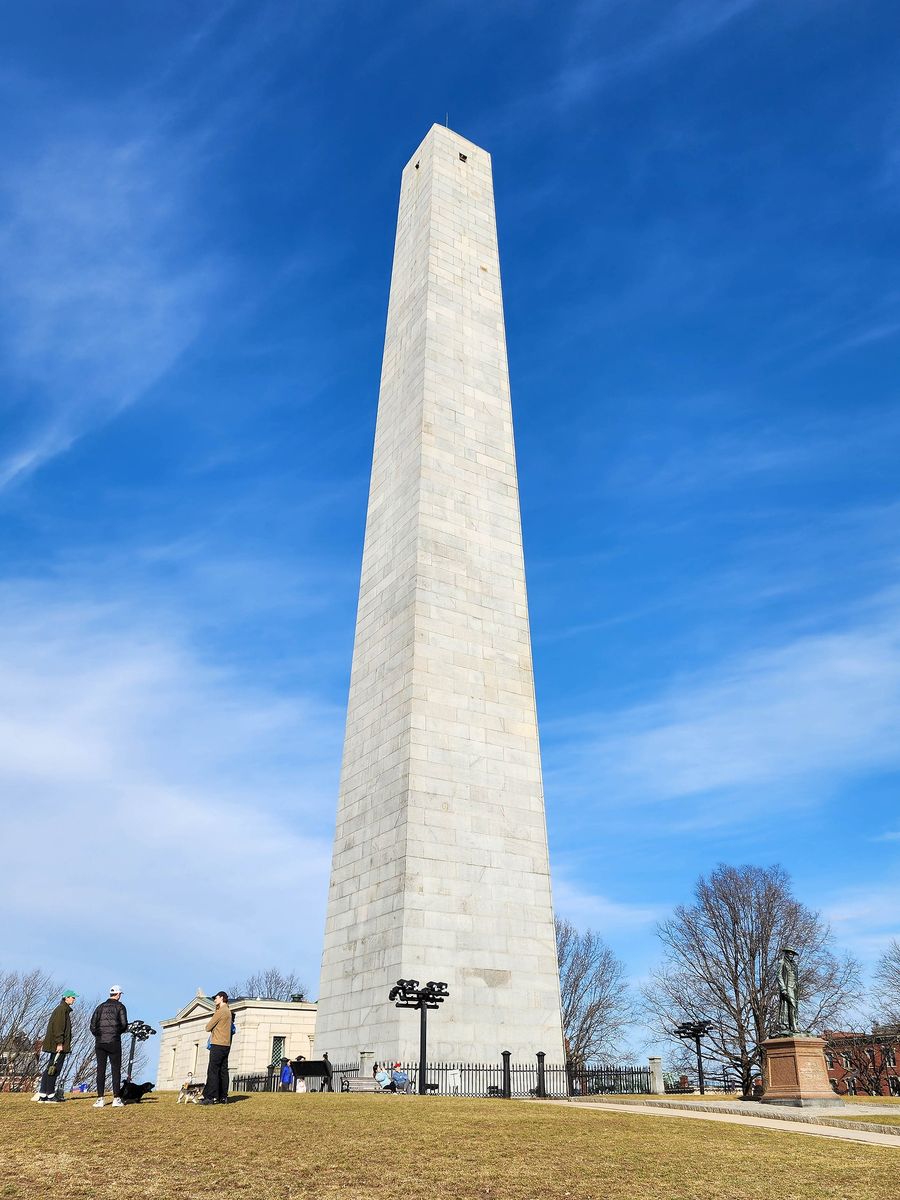 The 221-foot granite obelisk of the Bunker Hill Monument seen from below against a blue winter sky, with the lodge, the Colonel Prescott statue and a few visitors at its base.