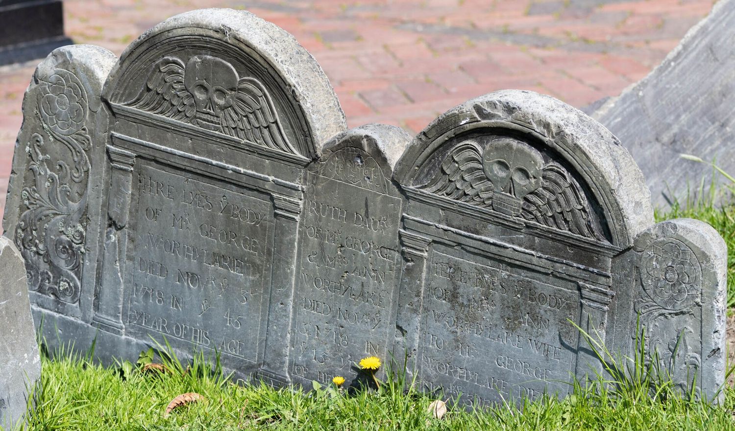 Three slate gravestones from 1718 at Copp's Hill Burying Ground, carved with winged skulls and rosettes, marking the graves of George Worthylake, his wife Ann and their daughter Ruth, with dandelions in the grass in front.