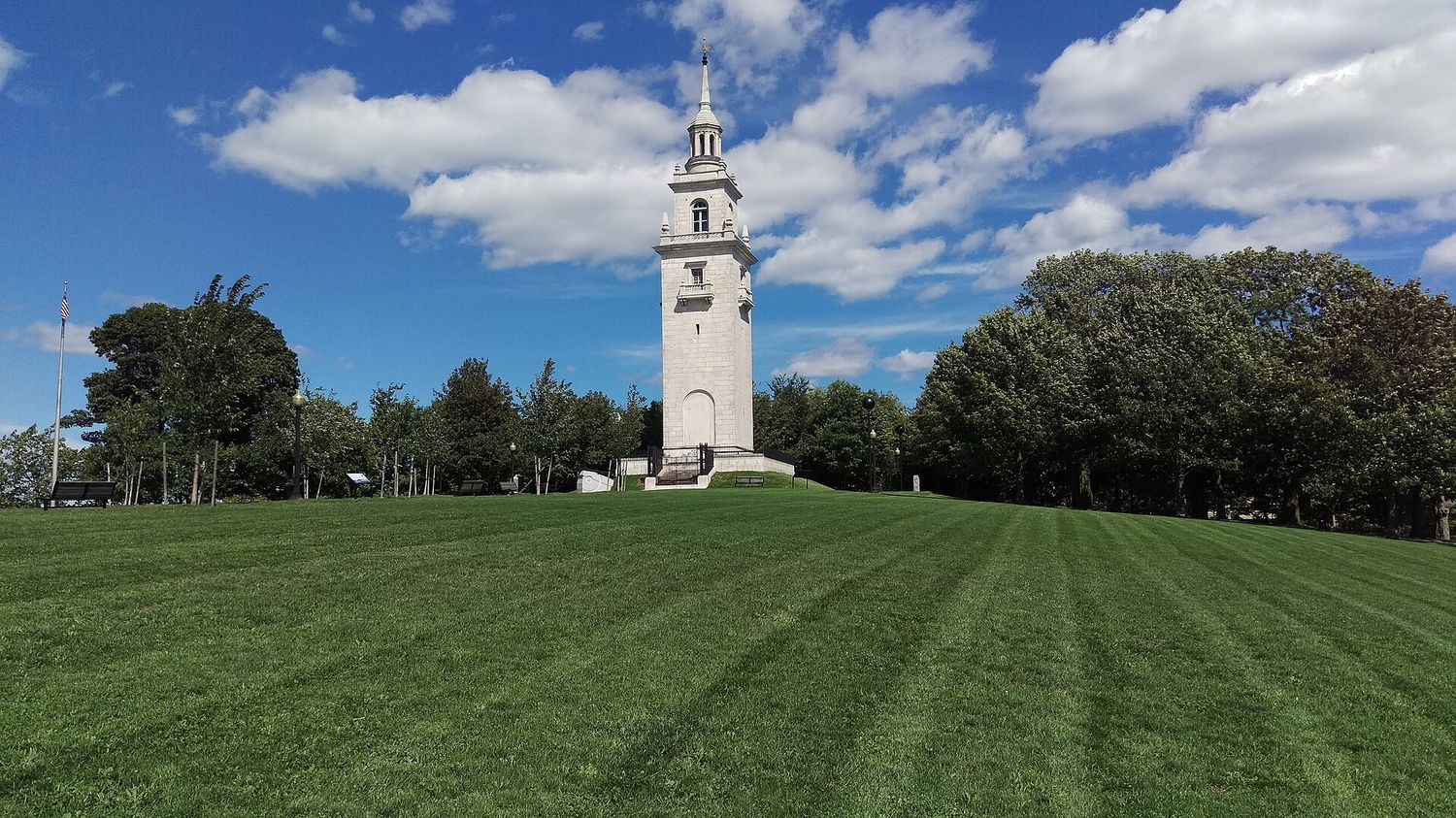 The white marble Dorchester Heights Monument, a tall Georgian Revival tower with a slender spire, standing on a green hilltop lawn in South Boston under a blue sky with white clouds.