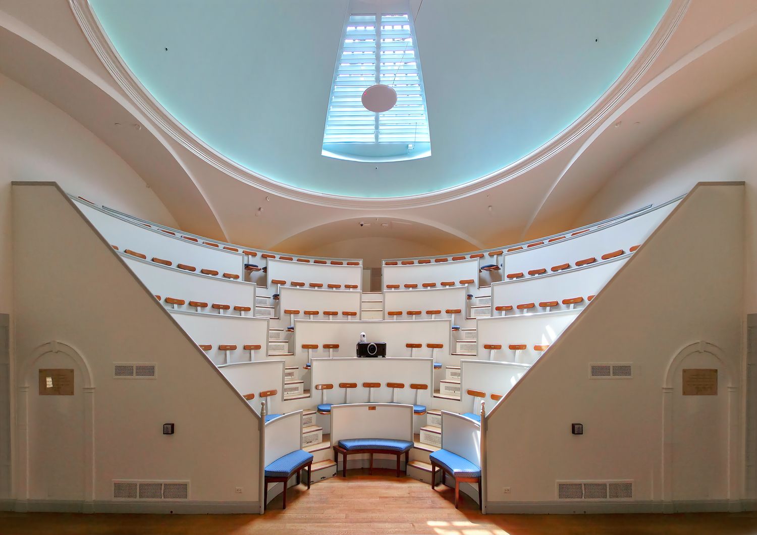 The interior of the Ether Dome at Massachusetts General Hospital, a white surgical amphitheater with steep curved tiers of wooden seats rising toward a pale domed ceiling and skylight.