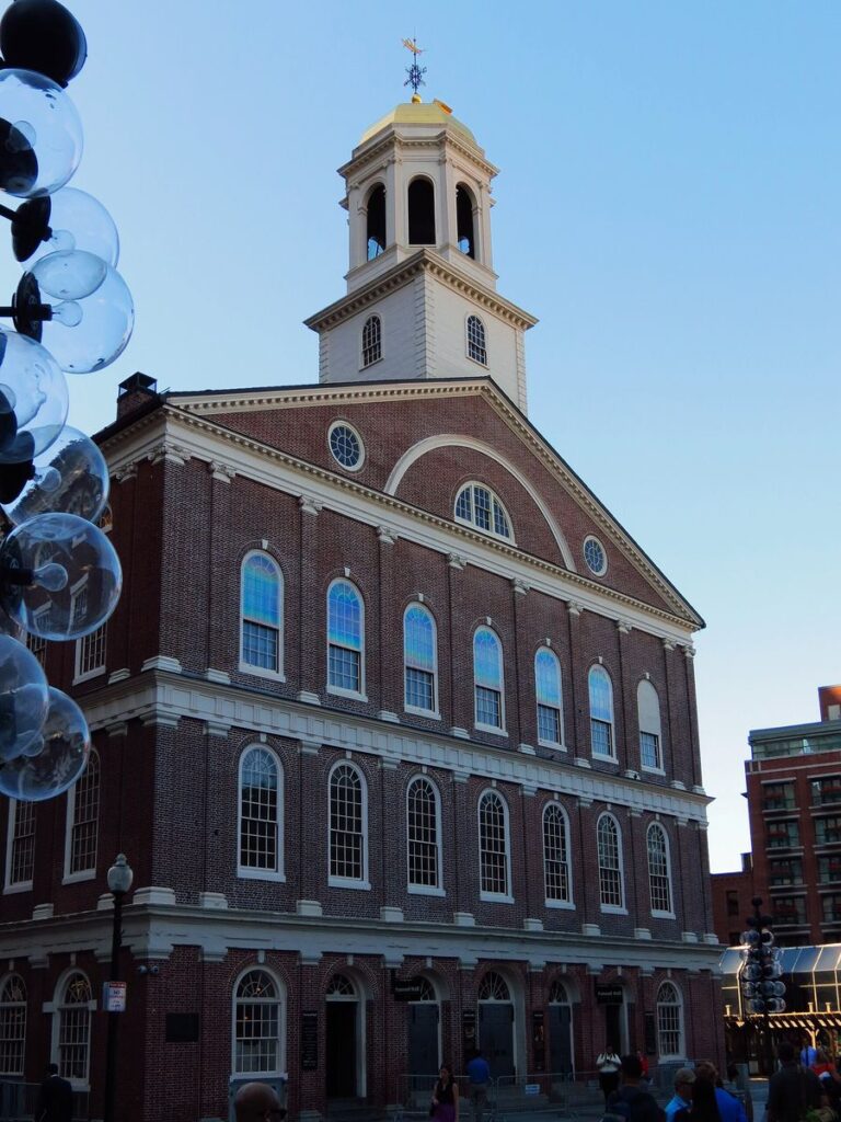 The red-brick facade of Faneuil Hall with its white cupola, gold dome and grasshopper weathervane against a clear blue evening sky, with visitors at the entrance below.