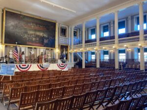 The Great Hall inside Faneuil Hall, Boston, with rows of wooden chairs, a large historical painting and a colonnaded gallery.