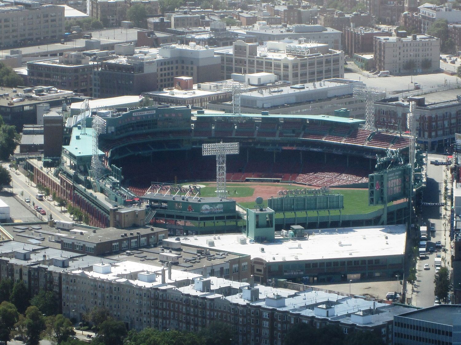 Fenway Park seen from high above, its green seats, light towers and diamond visible inside the ballpark, surrounded by the brick and concrete buildings of the Fenway neighborhood.