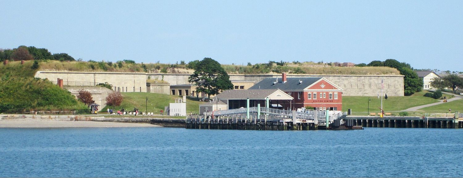 Georges Island seen from Boston Harbor, showing the long granite ramparts of Fort Warren above a grass slope, with the ferry pier and the red-brick visitor center at the water's edge.