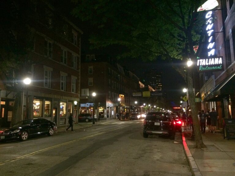 Hanover Street in Boston's North End at night, lined with lit restaurant signs and people on the sidewalk.