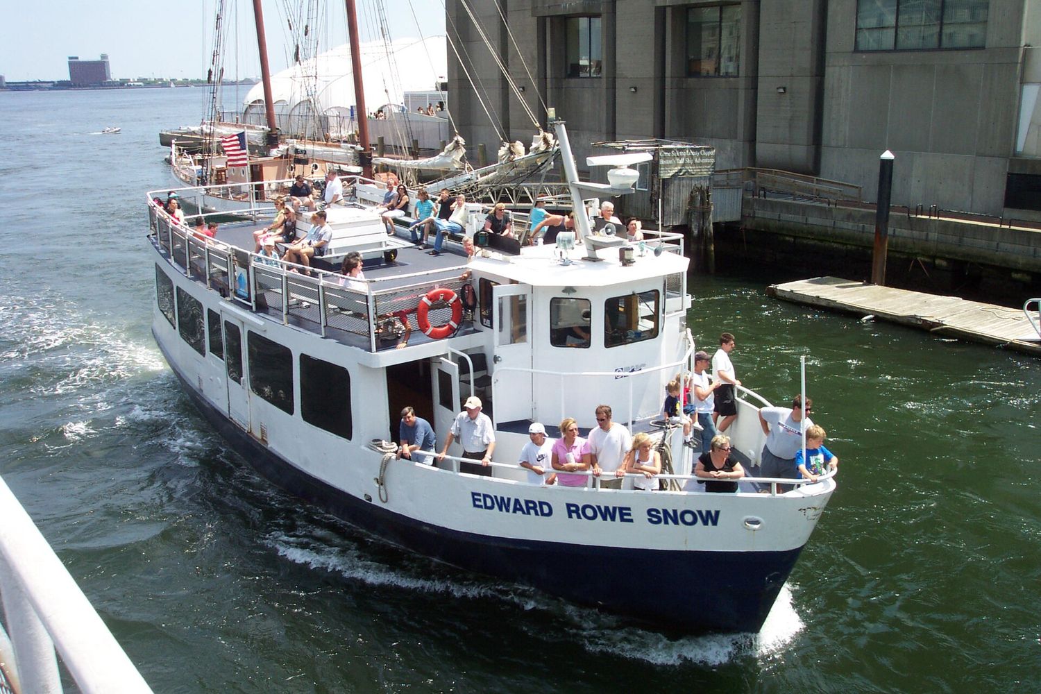 A white passenger boat named Edward Rowe Snow arriving at a Boston wharf with its open top deck full of passengers, a tall ship moored behind it.