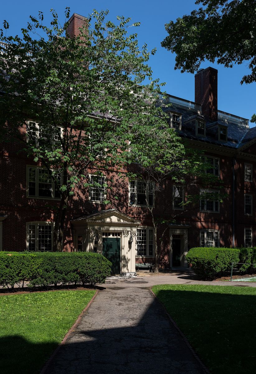 A brick Georgian building in Harvard Yard behind clipped hedges and a paved path, with trees in full leaf.
