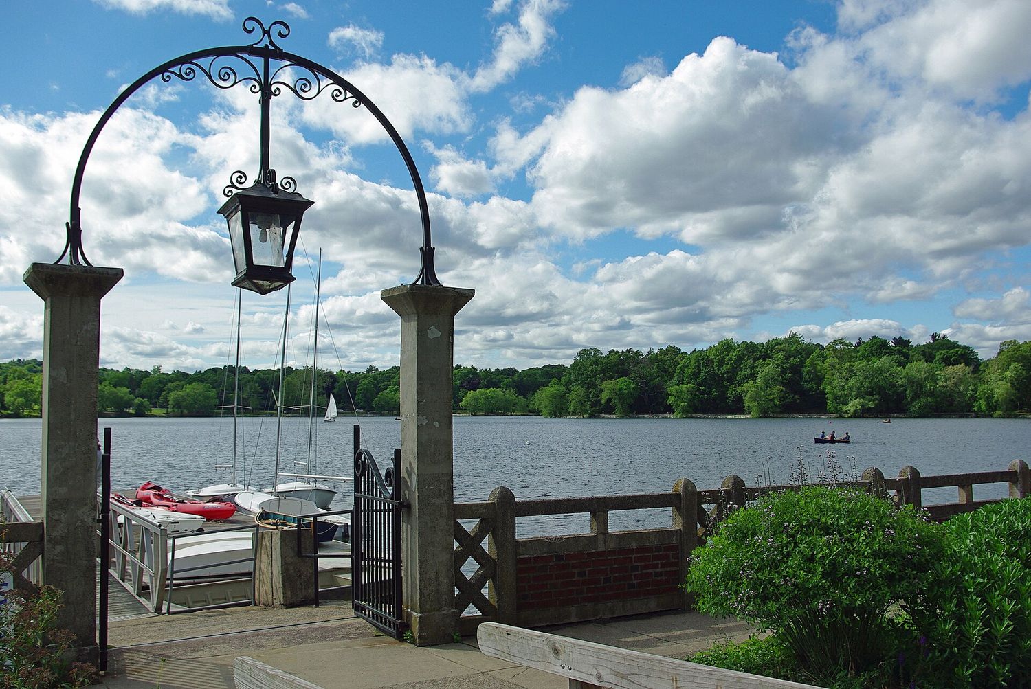 Jamaica Pond in Boston seen through an iron lamp arch, with a boathouse dock, moored sailboats and wooded banks beyond.