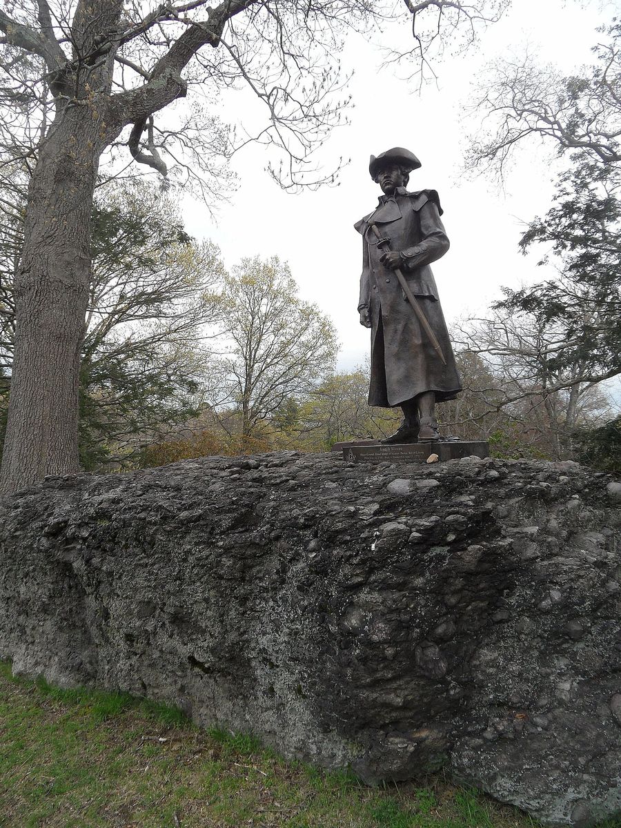 A bronze statue of the Revolutionary patriot Joseph Warren in a long coat and tricorn hat, holding a sword, standing on top of a large rough boulder among bare spring trees in Forest Hills Cemetery.