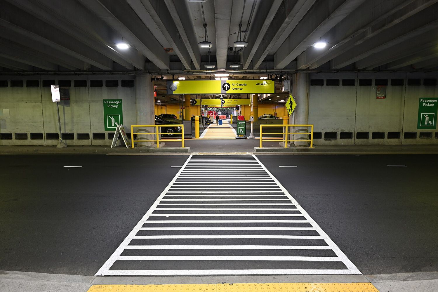 A wide crosswalk at Boston Logan Airport leading toward a Terminal B passenger pickup area under a concrete parking structure, with yellow overhead signage.