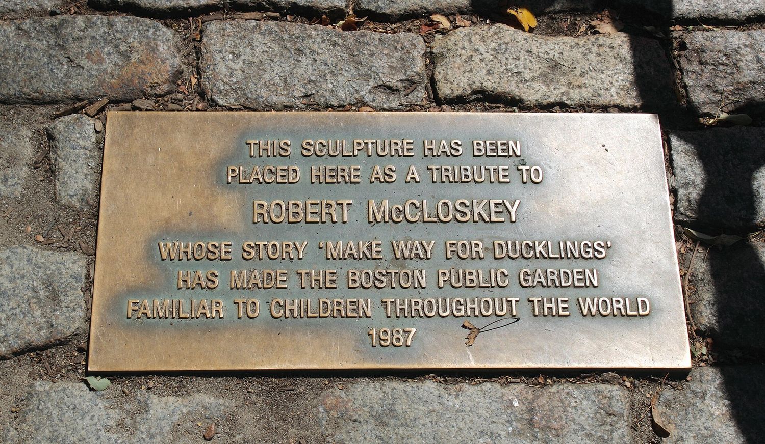 A bronze plaque set among granite cobbles reading: This sculpture has been placed here as a tribute to Robert McCloskey whose story Make Way for Ducklings has made the Boston Public Garden familiar to children throughout the world, 1987.