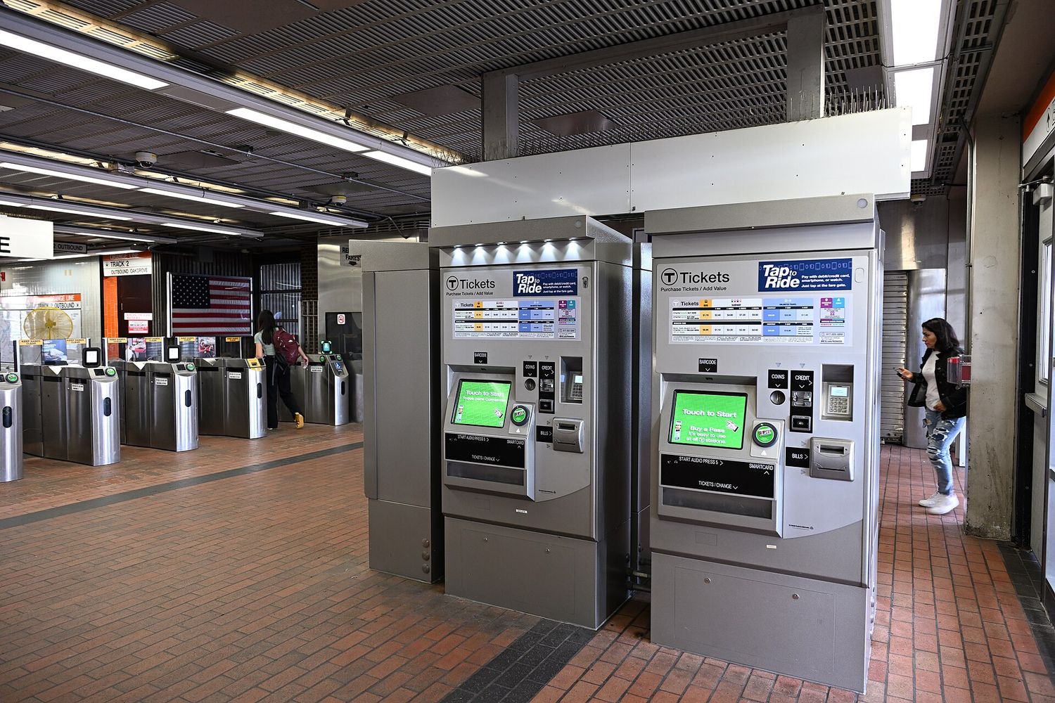 Two MBTA ticket vending machines with Tap to Ride signage in a subway station, with a row of fare gates and a passenger walking through in the background.