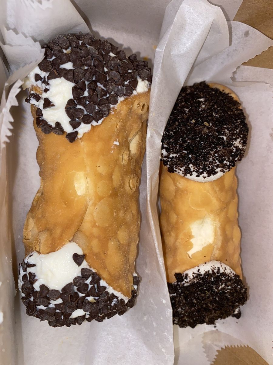 Two cannoli in a paper-lined box from Mike's Pastry, one rolled in chocolate chips and one in cookie crumbs.