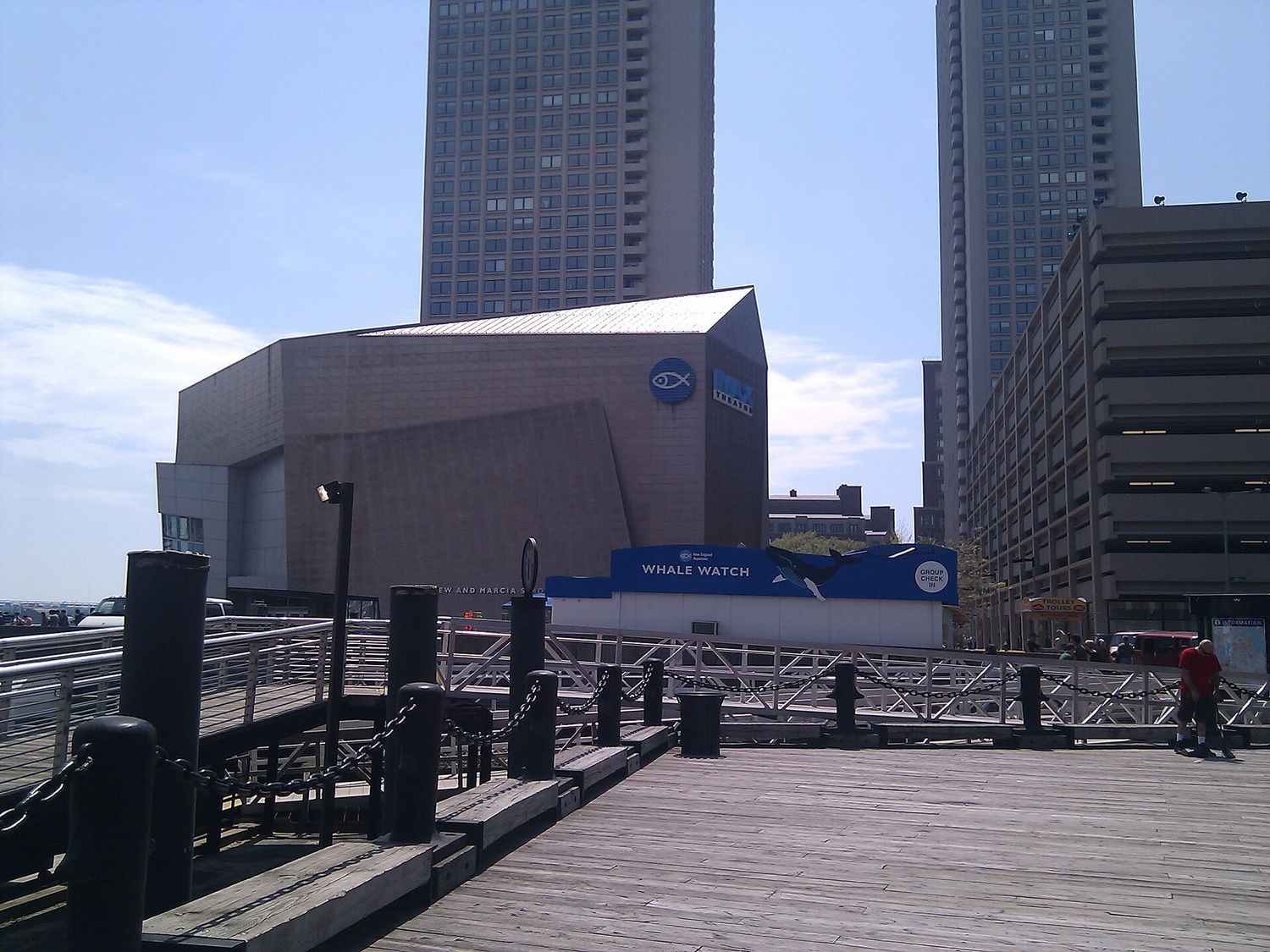The concrete New England Aquarium building and its IMAX theater seen from the wooden boardwalk of Central Wharf, with a blue Whale Watch ticket kiosk at the gangway.