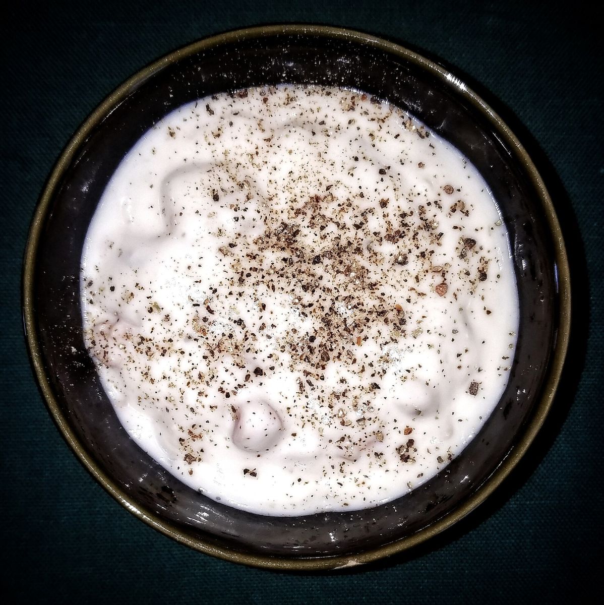 A dark ceramic bowl of thick white New England clam chowder, dusted with coarsely ground black pepper.