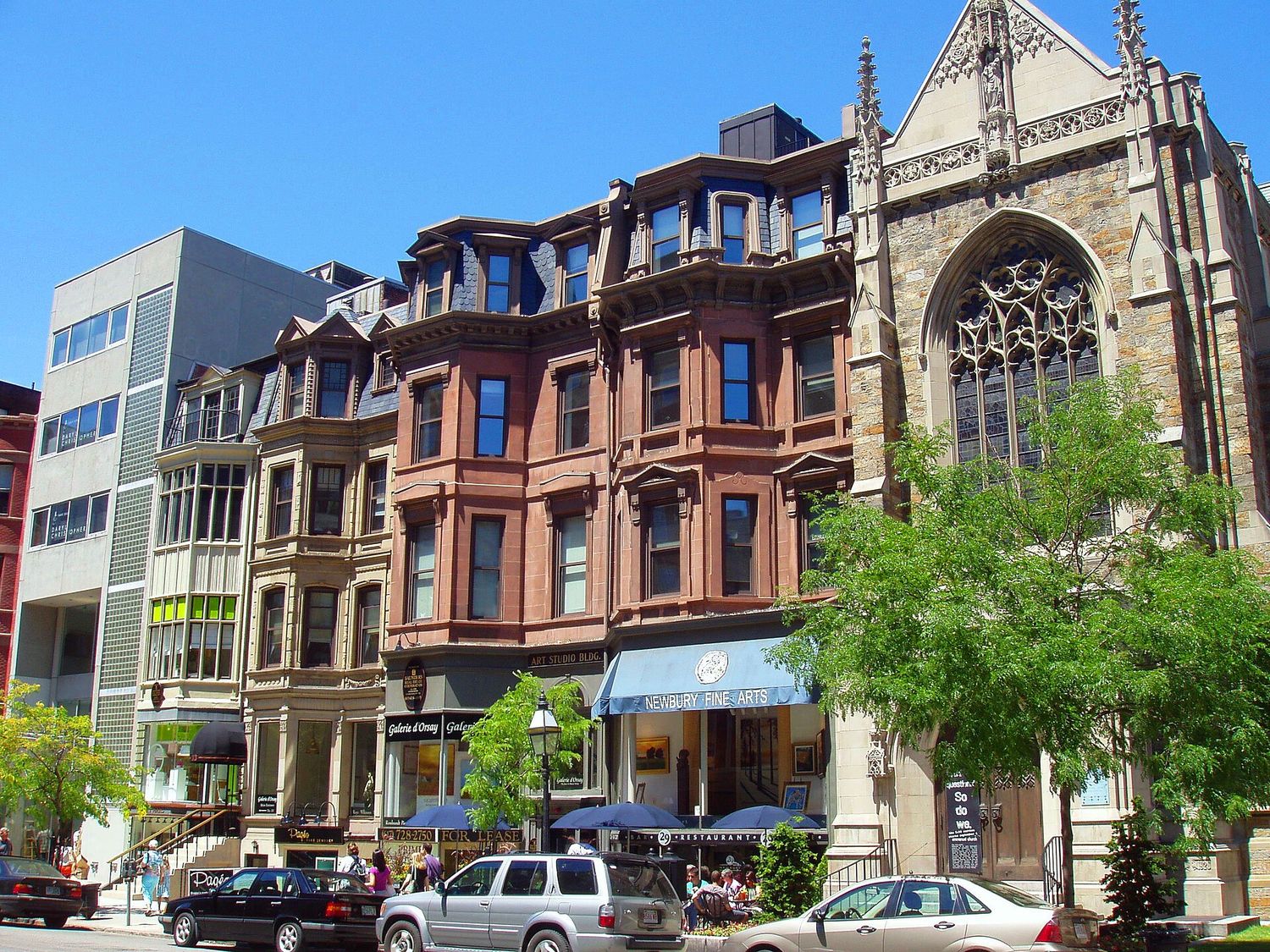 A row of Victorian brownstone buildings on Newbury Street in Boston, with gallery and restaurant frontages at street level and a Gothic stone church at the end of the block.