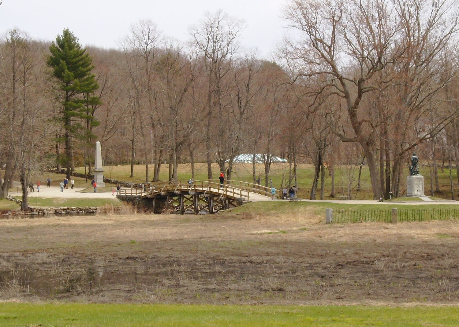 The wooden Old North Bridge over the Concord River, with the obelisk on one bank and the Minute Man statue on the other, and visitors walking across.