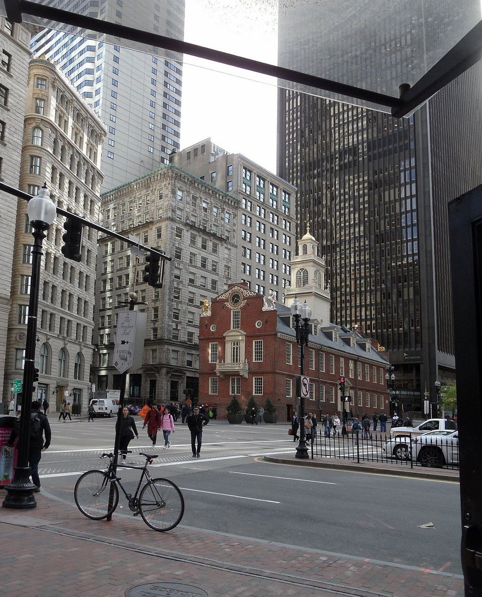 The brick Old State House with its gilded lion and unicorn and white cupola, standing at a downtown intersection surrounded on all sides by far taller glass and stone office towers, with a Freedom Trail signpost at the left.