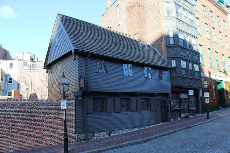 The dark gray clapboard Paul Revere House in the North End's North Square, with diamond-pane windows, beside taller brick buildings, and the red-brick Freedom Trail line running along the sidewalk in front.