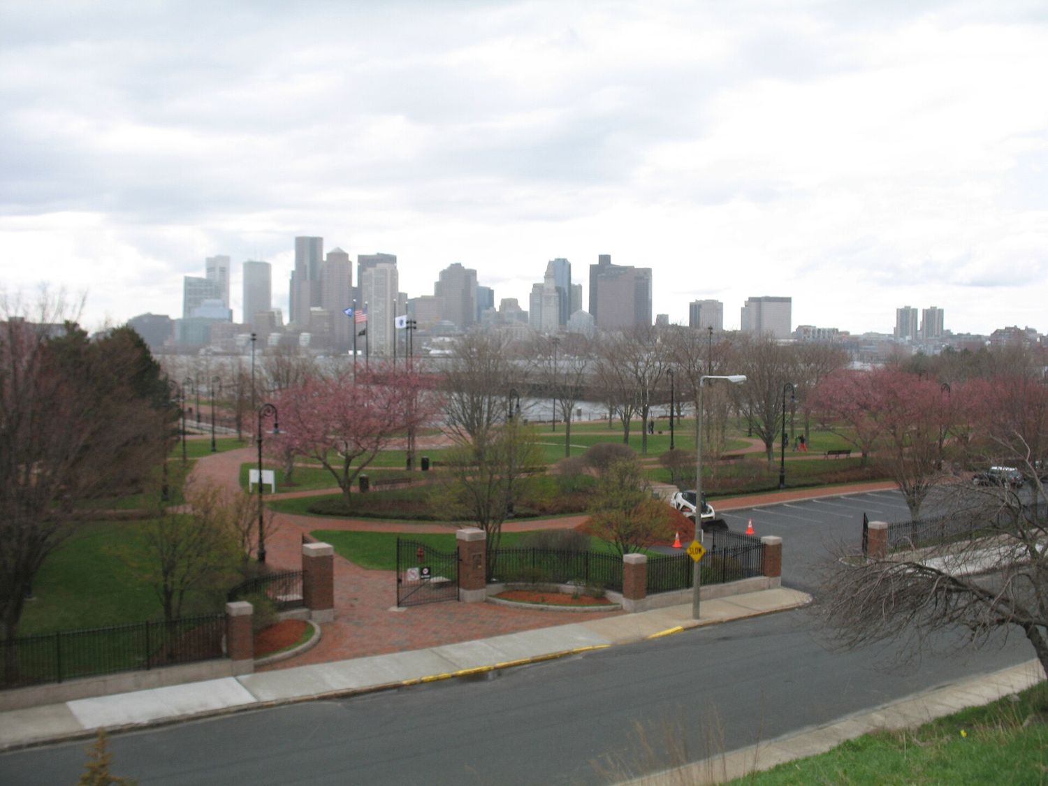 Piers Park in East Boston in spring, with brick paths, flowering trees and a flagpole leading to the water, and the downtown Boston skyline across the harbor under a cloudy sky.