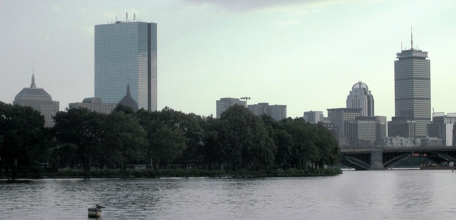 The Back Bay skyline across the Charles River, with the glass slab of 200 Clarendon at left and the Prudential Tower with its antenna at right, above a line of trees on the Esplanade.