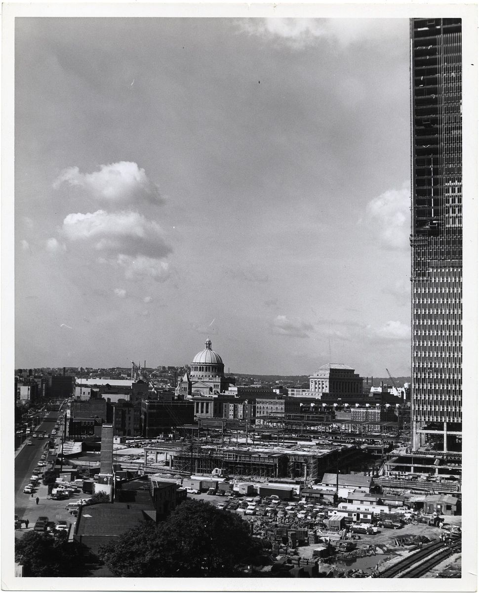 A black-and-white photograph of the Prudential Tower under construction, its steel frame rising at the right, with the dome of the Christian Science Mother Church and a wide construction site with parked cars in the foreground.