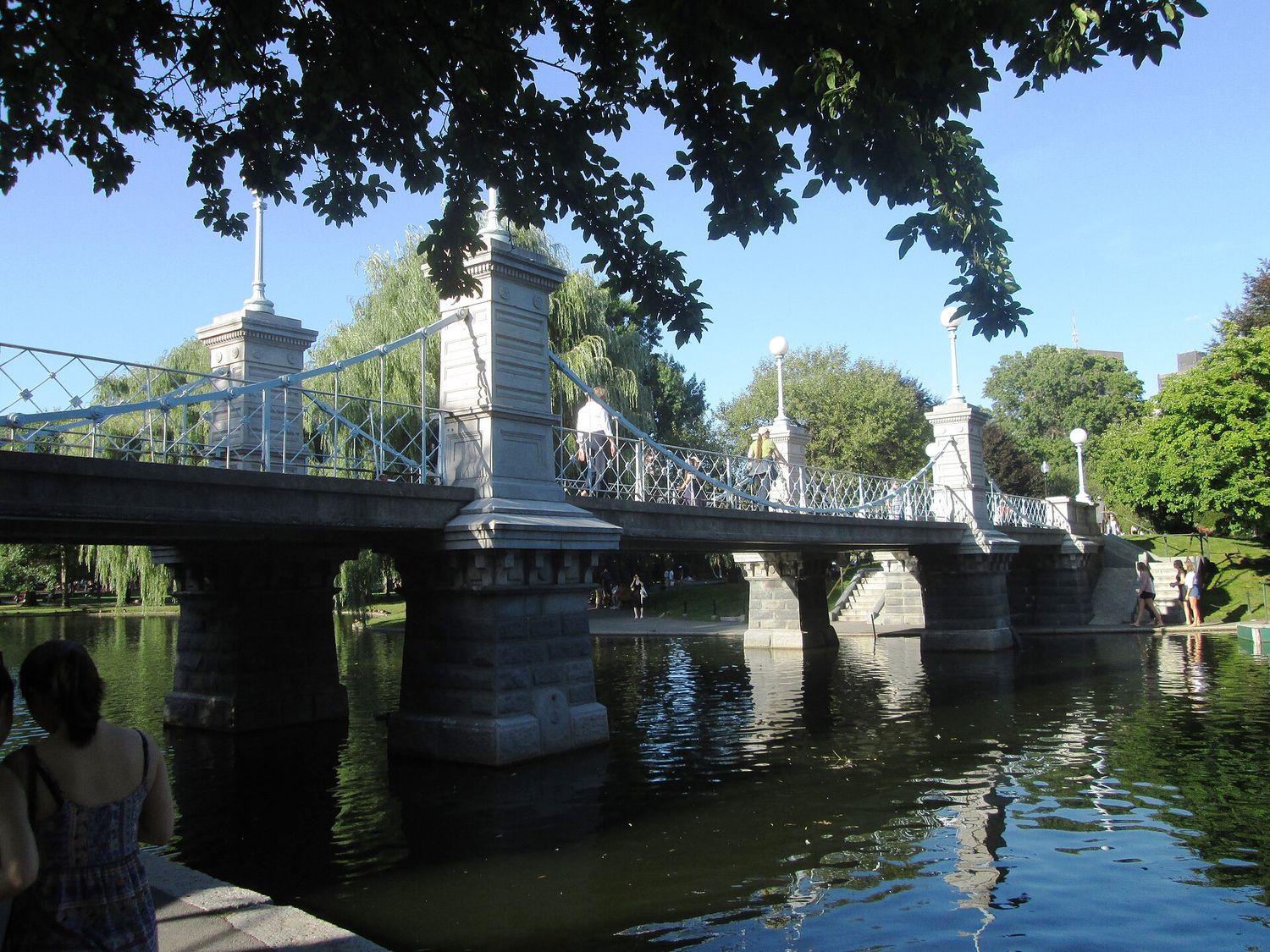 The granite-pier footbridge across the Public Garden lagoon, with pale blue decorative suspension cables and globe lamps on its posts, weeping willows behind and walkers crossing.