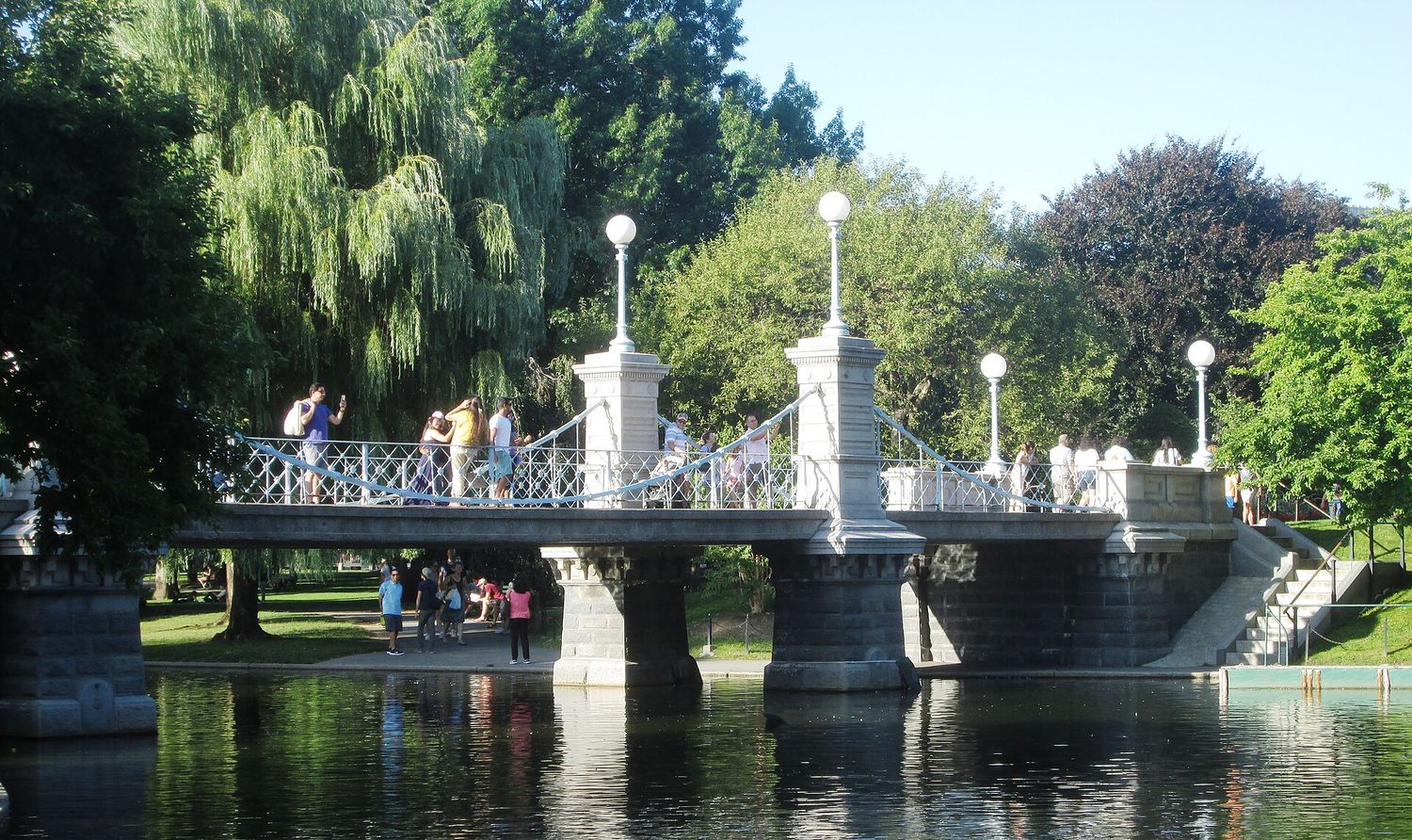 The small iron suspension bridge over the lagoon in Boston Public Garden, with white lamps, weeping willows behind it and the water reflecting the stonework.