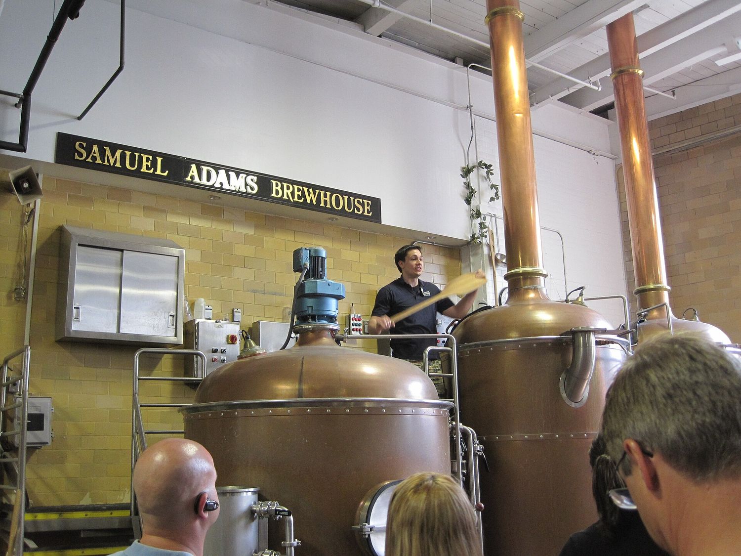 A guide with a wooden paddle standing behind two copper brew kettles beneath a Samuel Adams Brewhouse sign, addressing a group of visitors on a brewery tour.